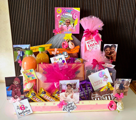 A 6th birthday gift basket filled with treats, toys, and party supplies, including a bottle of orange juice, chocolate bars, and baby photos, set against a dark wood slatted background.